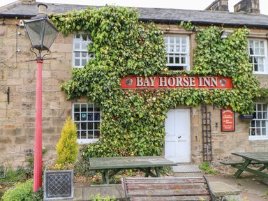 The exterior of a stone building with ivy climbing around windows and a sign that reads Bay Horse Inn with outdoor picnic tables and a red lamp post in front at The Garden Cottage in West Woodburn