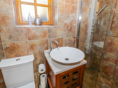A bathroom with a toilet, a round white sink on a wooden cabinet, a window with toiletries, and a glass shower at Bwthyn Carregwen in Dwygyfylchi