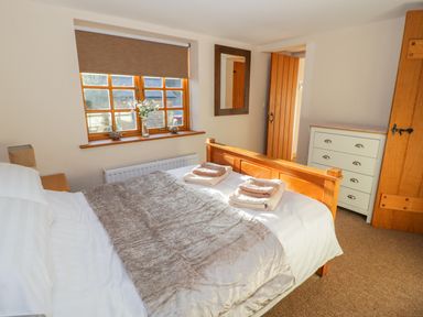 A bedroom with a wooden bed, folded towels on the bed, a window with a blind, a mirror on the wall, and a chest of drawers at Bwthyn Carregwen in Dwygyfylchi