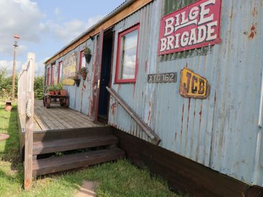 An exterior view of a weathered metal-sided cabin with wooden steps and potted plants at Crows Cabin in Gloucester