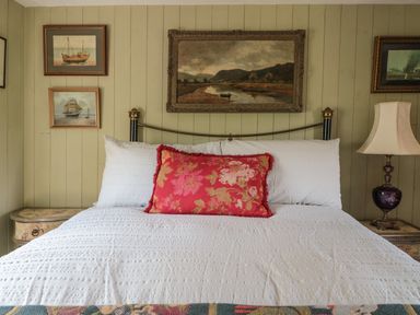 A bed with white pillows and a red floral cushion with paintings on the wall and a lamp on a bedside table at Crows Cabin in Gloucester