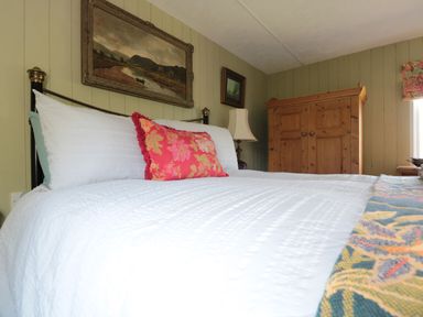 A bedroom with a white bedspread a red decorative pillow a wooden wardrobe and paintings on paneled walls at Crows Cabin in Gloucester