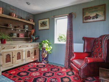 A room with a red leather armchair a wooden cabinet with shelves and copper kettles a window with a curtain and a red patterned rug on the floor at Crows Cabin in Gloucester