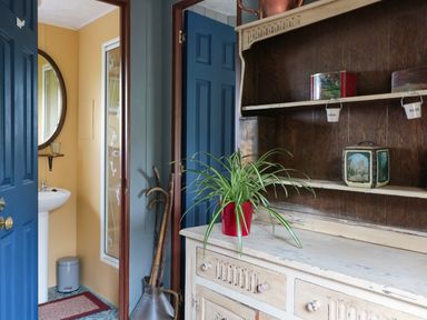 An entryway with open blue doors leading to a bathroom with a sink and round mirror and a wooden shelf unit with a potted plant at Crows Cabin in Gloucester