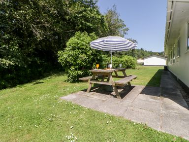 An outdoor picnic table with a striped umbrella and glasses on a grassy area beside buildings at Tranquillity Rosecraddoc Lodge near Liskeard