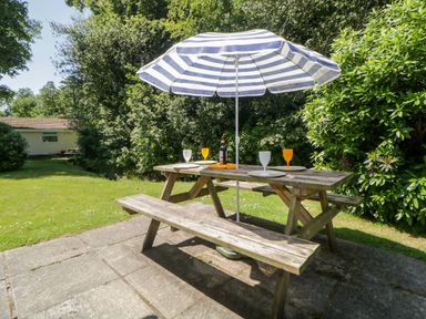 A wooden picnic table with plates and glasses under a striped umbrella in a garden at Tranquillity Rosecraddoc Lodge near Liskeard