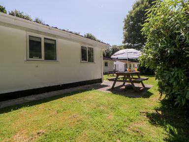A picnic table with an umbrella and drinks on a lawn next to white buildings surrounded by trees at Tranquillity Rosecraddoc Lodge near Liskeard