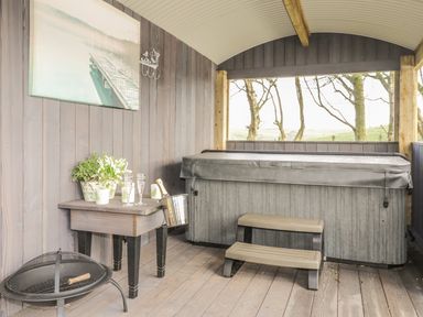 A covered outdoor area with a hot tub a small table with plants champagne glasses and a champagne bucket and a fire pit at High Rigg Shepherd's Delight in Brampton Cumbria