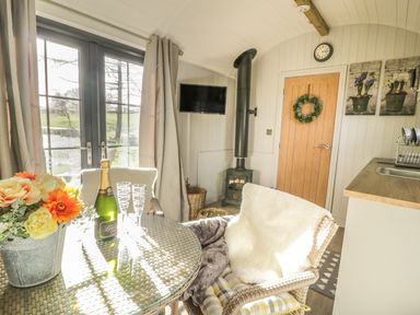 A dining area with a glass table a bottle of champagne and flowers near a window and a wood stove at High Rigg Shepherd's Delight in Brampton Cumbria