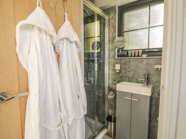 A bathroom with two white robes hanging on the door a shower with glass doors a sink with a cabinet below and toiletries on the window sill at High Rigg Shepherd's Delight in Brampton Cumbria