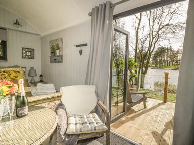 A small room with a wicker chair, glass table with a champagne bottle and glasses, a bed with pillows and folded towels, an open door leading to a wooden deck overlooking a lake at High Rigg Shepherd's Delight in Brampton Cumbria
