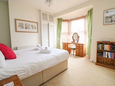A bedroom with a bed, wooden dresser, bay window with green curtains, bookshelf, and framed floral pictures at Brewers House in Weymouth