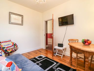 A living room with a blue sofa colorful pillows a black patterned rug a wooden dining table with chairs a fruit bowl a wall-mounted TV a small white table with a router and an open wooden door at Brewers House in Weymouth