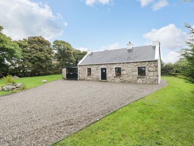A stone cottage with a gravel driveway and green lawn surrounded by trees at An Teach Ban in Rossaveel County Galway