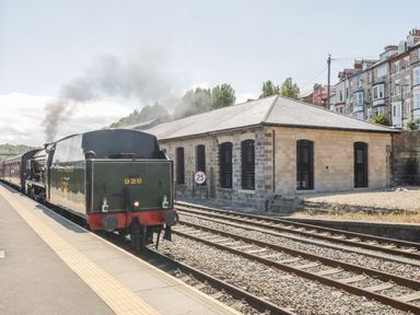 A steam train at a railway platform next to tracks and a stone building at Evening Star @ Engine Shed in Whitby