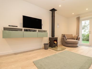 A living room with a wall-mounted television a wood-burning stove a round armchair and a glass door at Kober House in Mullion
