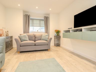 A living room with a grey sofa blue cushions a window with curtains a wall-mounted TV and a small side table with flowers at Kober House in Mullion