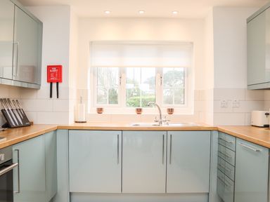 A kitchen with blue cabinets wooden countertops a sink under a window and a fire blanket on the wall at Kober House in Mullion