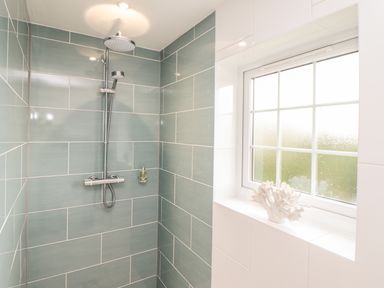 A shower area with green tiles a chrome showerhead and a window with frosted glass at Kober House in Mullion