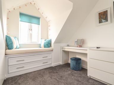 A small room with a window seat with cushions a desk a vanity stool and a framed picture on the wall at Kober House in Mullion