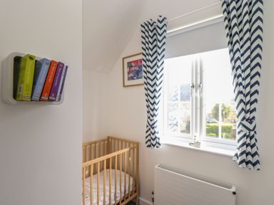 A small room with a wooden crib white bedding a window with blue and white chevron curtains a radiator and a small shelf with books at Kober House in Mullion