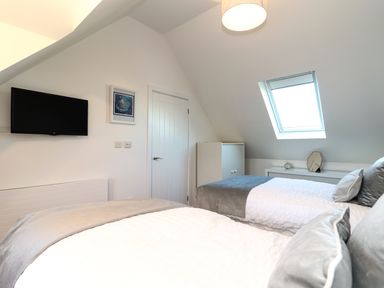 A bedroom with two single beds a wall-mounted TV a door a white cupboard under a skylight window and a bedside table with a clock and mirror at Kober House in Mullion