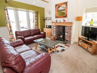 A living room with two red leather sofas a coffee table a brick fireplace with a wood stove a wooden TV stand with a television and patterned curtains at Hemmel Cottage in Chatton