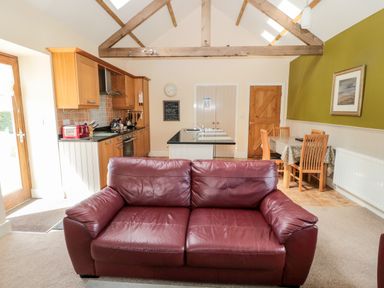 An open living space with a red leather sofa kitchen area with wooden cabinets and a black countertop and dining table with wooden chairs at Hemmel Cottage in Chatton