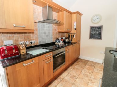 A kitchen with wooden cabinets black countertops tiled floor and walls a red toaster a knife block and a wall clock with a board at Hemmel Cottage in Chatton
