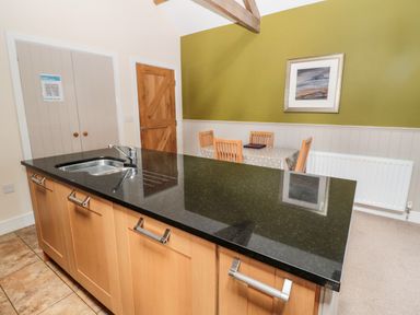 A kitchen island with sink and wooden cabinets next to a dining table with four chairs and a framed picture on a green wall at Hemmel Cottage in Chatton