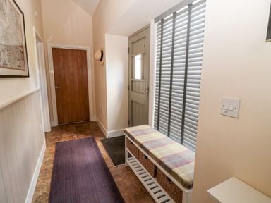 An entryway with a wooden door a green wall door blinds a bench with cushions and baskets and a purple rug on tiled floor at Hemmel Cottage in Chatton