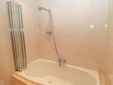 A bathtub with a showerhead and folding glass shower screen in a tiled bathroom at Hemmel Cottage in Chatton