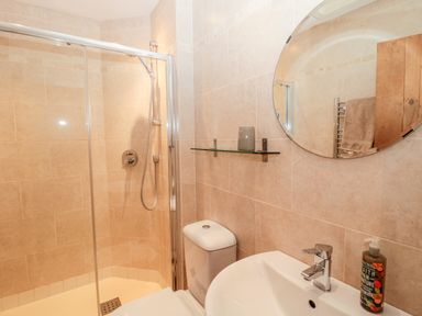 A bathroom with a shower enclosure a toilet a sink with a soap dispenser a round mirror and a glass shelf at Hemmel Cottage in Chatton