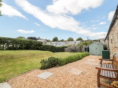 A garden with a gravel path stone slabs a wooden bench green shed and lawn at Hemmel Cottage in Chatton