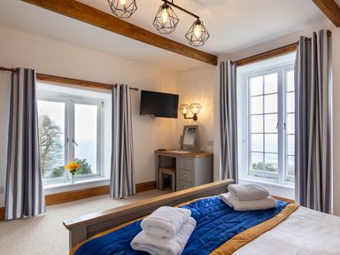 A bedroom with a bed covered with a blue and yellow blanket with folded towels on top a desk with a mirror a wall mounted television two large windows with blue and white striped curtains and a vase with yellow flowers on a windowsill at Bayview Terrace in Lynton