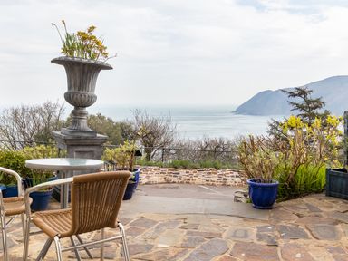 An outdoor patio with a round table and two wicker chairs overlooking the sea and hills with potted plants and a large decorative urn at Bayview Terrace in Lynton