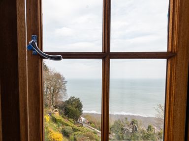 A view through a wooden window showing trees bushes a pebble beach and the ocean at Bayview Terrace in Lynton