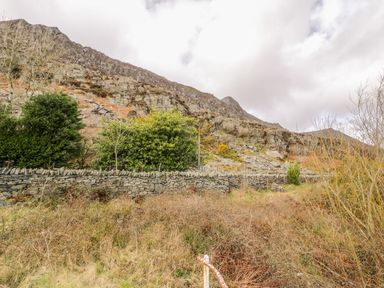 A stone wall with bushes and trees in front of a rocky hillside under a cloudy sky at Horeb Chapel House in Tanygrisiau