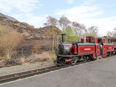 A steam train on railway tracks next to a paved platform with hills and trees in the background at Horeb Chapel House in Tanygrisiau