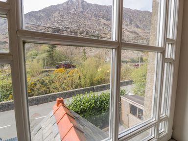 View through a window showing a mountain a road a stone wall shrubs a small wooden shed and a red train at Horeb Chapel House in Tanygrisiau