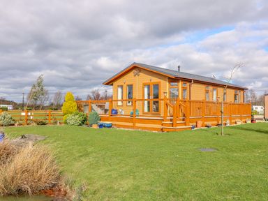A wooden lodge with a deck and railing on a grassy lawn with shrubs and a small tree at Lake View Lodge in Marston near Barkston