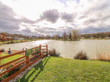 A fenced grassy area by a lake with houses and trees in the background at Lake View Lodge in Marston near Barkston