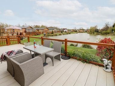 An outdoor deck with wicker furniture and a glass table overlooking a lake with houses in the background at Lake View Lodge in Marston near Barkston