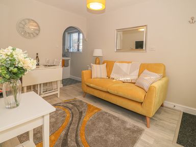 A living room with a yellow sofa a rug a small white table with flowers and an archway to a kitchen at Holly Cottage in Criccieth