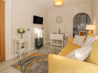 A living room with a yellow sofa a small dining table a wall-mounted tv and a doorway to a kitchen at Holly Cottage in Criccieth