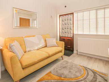 A yellow sofa with patterned pillows and a blanket in a room with a wooden door with a sailboat curtain and a window with blinds at Holly Cottage in Criccieth