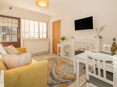 A small living room with a yellow sofa a dining table with chairs a wall-mounted tv and a window with blinds at Holly Cottage in Criccieth