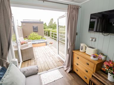 A living area with a sofa pillow and a dresser with a kettle toaster and snacks facing a mounted television and glass doors opening to an outdoor deck with chairs hot tub and small shed at Hydrangea in Bretforton