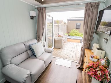 A small living room with a gray sofa two pillows a wooden cabinet with a toaster kettle and flowers and an open door leading to a wooden deck with chairs and a hot tub at Hydrangea in Bretforton