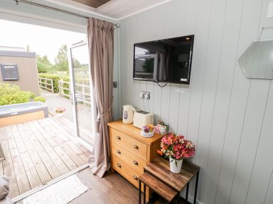A corner of a room with a chest of drawers a wooden table with flowers a wall mounted TV and an open glass door leading outside at Hydrangea in Bretforton